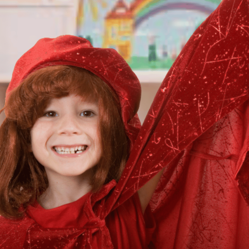 Smiling elementary school girl wears wizard costume with maroon floppy hat, wig and sparkle cape.